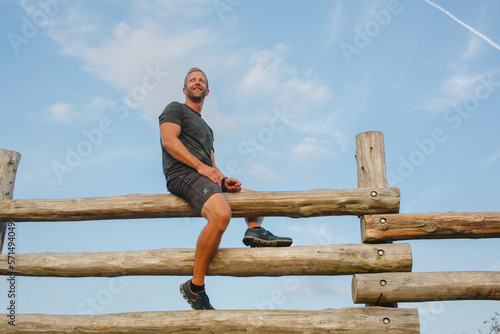 A proud smiling man sits high atop a tall wood fence against blue sky photo