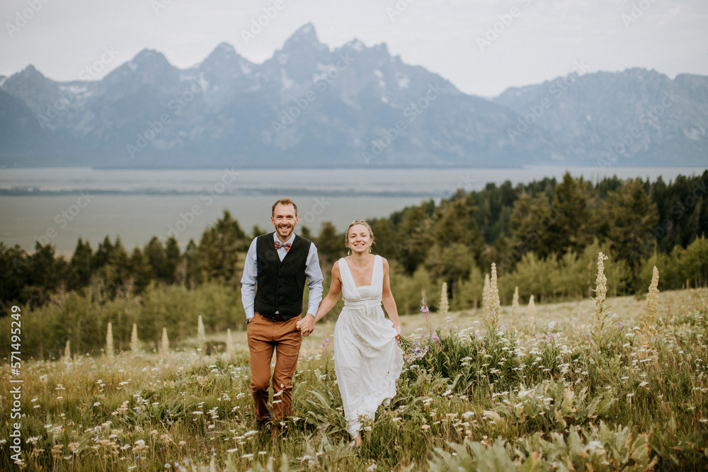 happy smiling newlyweds walk through field of wildflowers in Tetons happy smiling newlyweds walk through field of wildflowers in Tetons