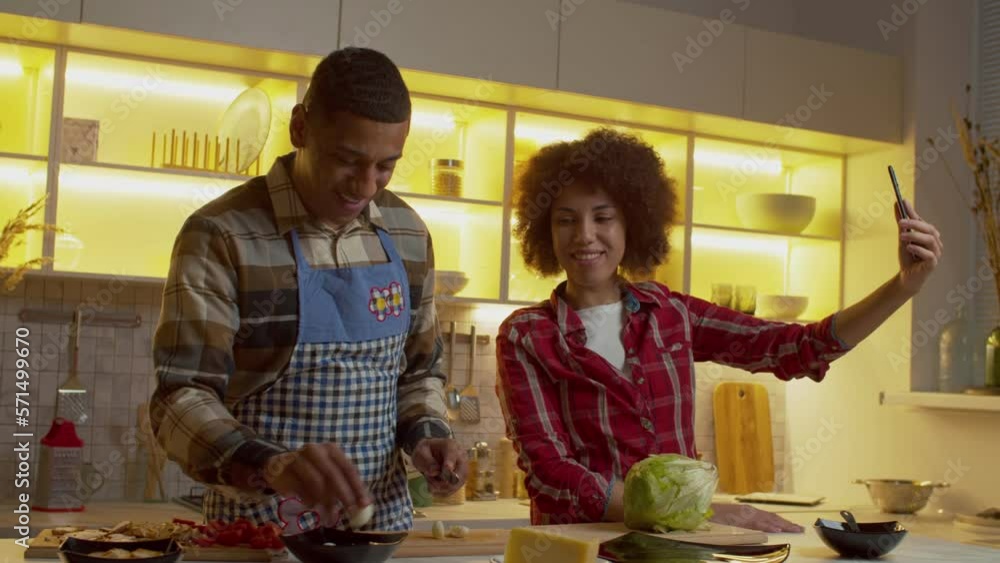 Cheerful proud pretty African American woman recording video content for social media on smartphone, sharing happy moments of family life while positive black man preparing dinner in kitchen. Cheerful proud pretty African American woman recording video content for social media on smartphone, sharing happy moments of family life while positive black man preparing dinner in kitchen.