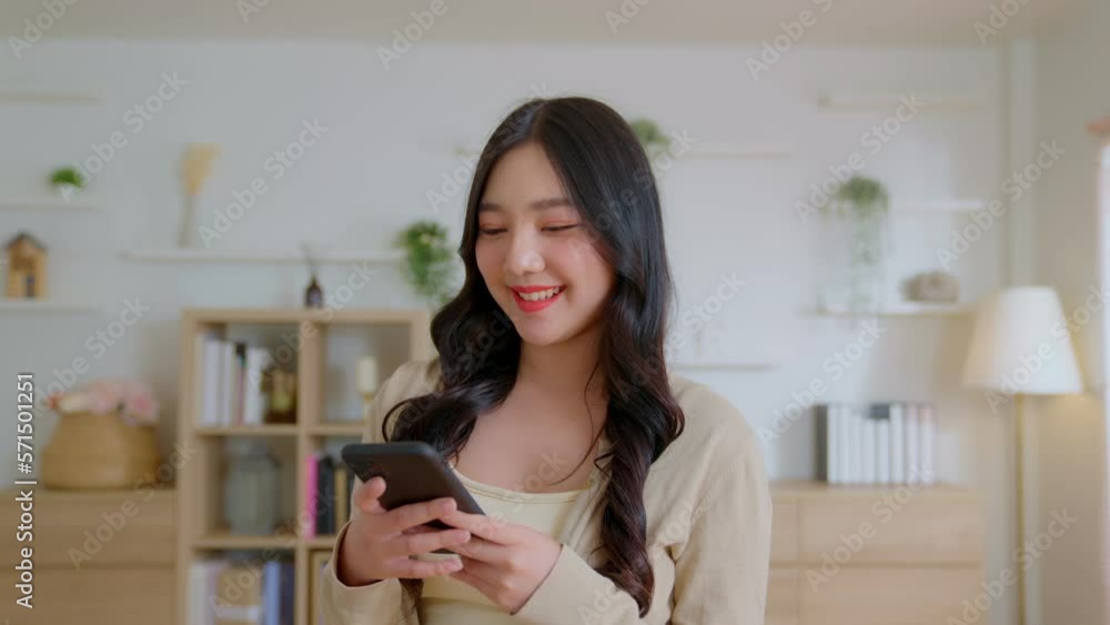 Attractive beautiful Asian woman happy playing with her mobile phone in the living room of new home. Female looks at the camera and smiles happily and very cute. Asia girl close up front portrait Attractive beautiful Asian woman happy playing with her mobile phone in the living room of new home. Female looks at the camera and smiles happily and very cute. Asia girl close up front portrait