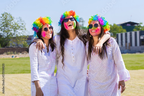 Group of playful young indian female friends wearing white kurta and colorful rainbow hair wigs celebrating holi festival outdoor at park.