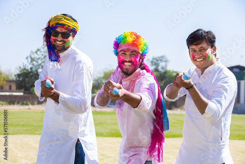Cheerful young indian male friends wearing white kurta outfit playing with pichkari celebrating holi festival outdoor at park.