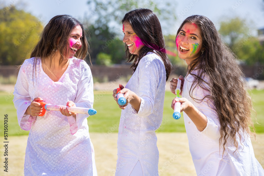Obraz premium Group of young indian girl friends wearing white kurta outfit playing with pichkari celebrating holi festival outdoor at park.