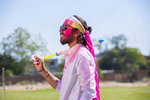 Young indian man with sunglasses holding pichkari in hand play holi festival with his face painted with powder color or gulal.