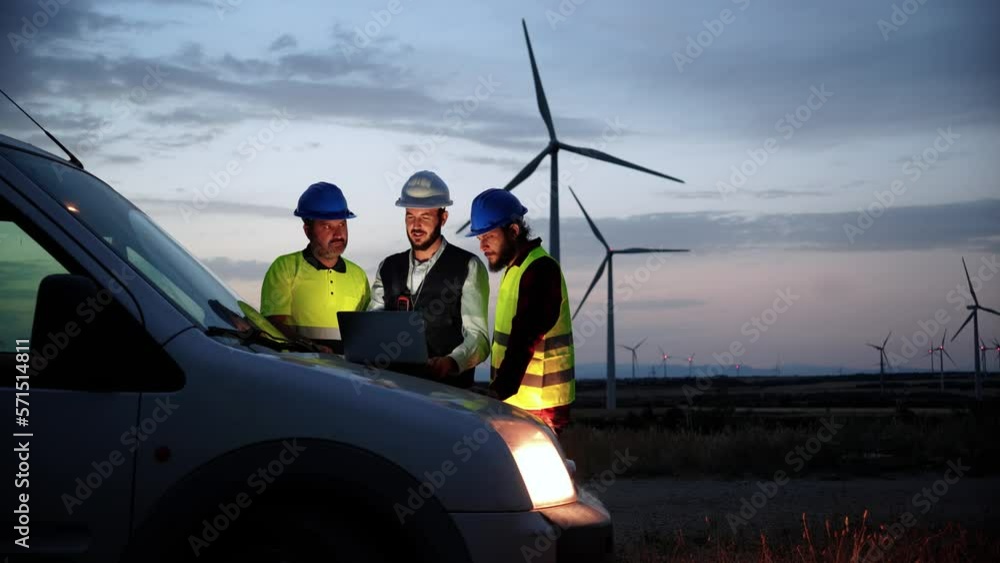 Team work of three renewable energy engineers with helmets and protection equipment, working and planing at night with laptop computer on top of the car with a wind power generators in the background Team work of three renewable energy engineers with helmets and protection equipment, working and planing at night with laptop computer on top of the car with a wind power generators in the background