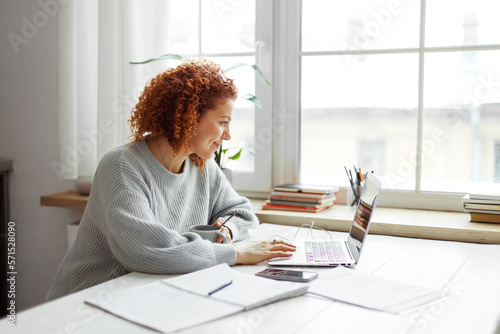Side view of pretty young redhead teacher sitting at table next to huge window at university, drinking coffee and checking e-mail during brake, having rest between lessons, preparing material
