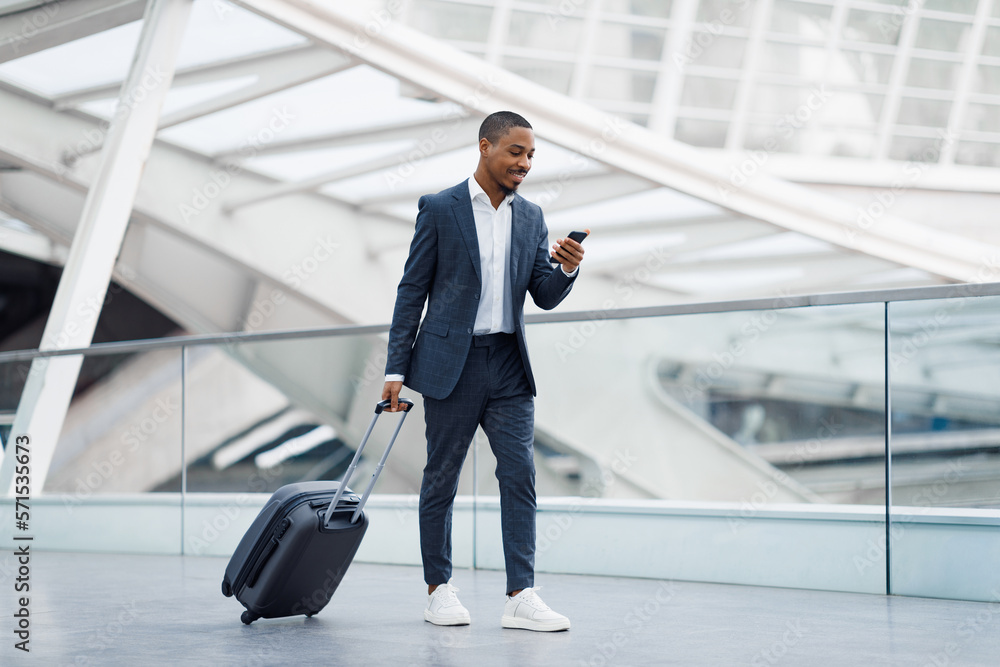 Handsome Black Businessman In Airport Walking With Suitcase And Using Smartphone Handsome Black Businessman In Airport Walking With Suitcase And Using Smartphone