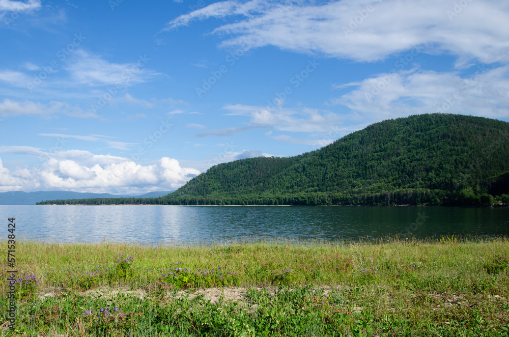 Bay on Lake Baikal in sunny summer weather. In the smooth water, clouds and mountains covered with forest are reflected.
