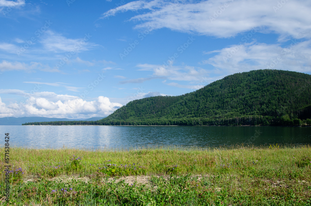 Bay on Lake Baikal in sunny summer weather. In the smooth water, clouds and mountains covered with forest are reflected. Bay on Lake Baikal in sunny summer weather. In the smooth water, clouds and mountains covered with forest are reflected.