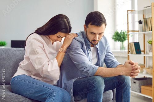 Young woman trying to apologize and make peace with her husband after quarrel sitting on sofa at home. Sad female calming offended man. Relationship problems, conflicts, quarrel and divorce concept. photo