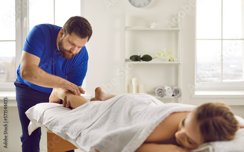 Male therapist work with female client lying on table in wellness salon or spa. Masseur or physiotherapist do manual massage patient leg relive strain or spasm in muscles. Relaxation and recovery.