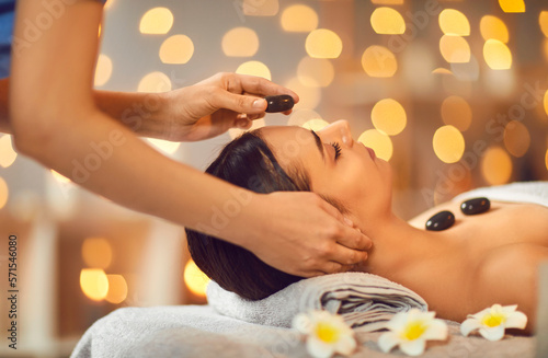 Close up portrait of a pretty young brunette woman with closed eyes lying relaxing in spa salon getting face and head massage therapy with hot stones. Wellness and beauty day concept.