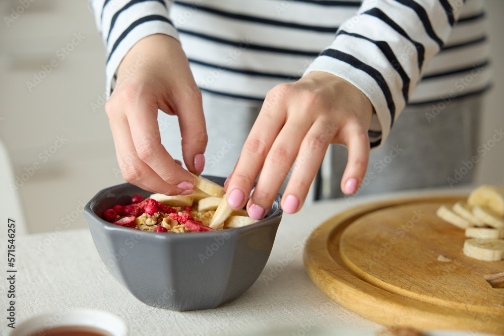Cropped shot of unrecognizable woman with hands decorating putting bananas on bowl with strawberries smoothie and granola for breakfast on kitchen countertop at home. Healthy lifestyle concept.