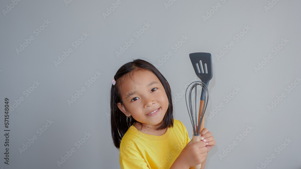 Happy asian toddler girl holding kitchen utensils over grey background. Cooking education concept for children. Happy asian toddler girl holding kitchen utensils over grey background. Cooking education concept for children.