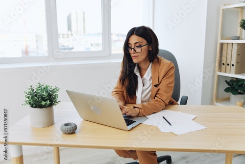 Business woman working in the office at her desk with a laptop in a beige suit and white shirt with glasses, work in the office writing a message