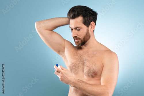Shirtless handsome man spraying deodorant under arm for fragrance, hygiene and fresh scent, blue background
