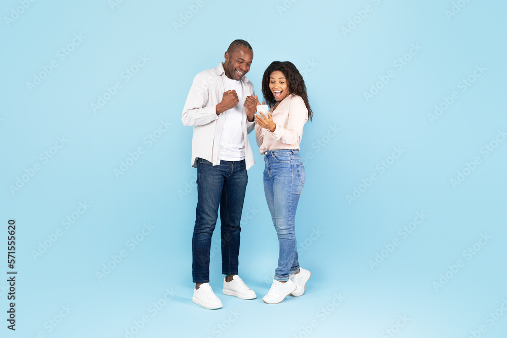 Overjoyed black couple holding smartphone and shaking fists in joy and excitement, blue studio background Overjoyed black couple holding smartphone and shaking fists in joy and excitement, blue studio background
