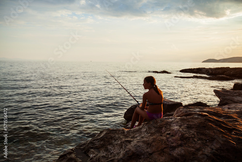  Child girl have fun on the sea rocky shoreline fishing at sunset light. Summer travel holiday. Beautiful summer scene. 