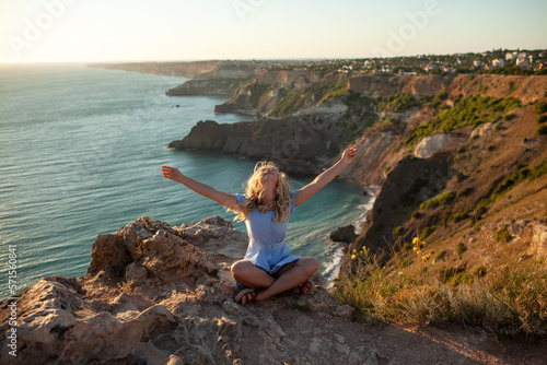 The traveller's girl with her hands raised sit and admires the nature of the rocks, sun and water. the tourist woman is dressed in summer clothes, blue sundress.