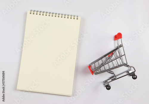flat lay of blank page opened notebook and shopping trolley or shopping cart on white background. shopping list concept.