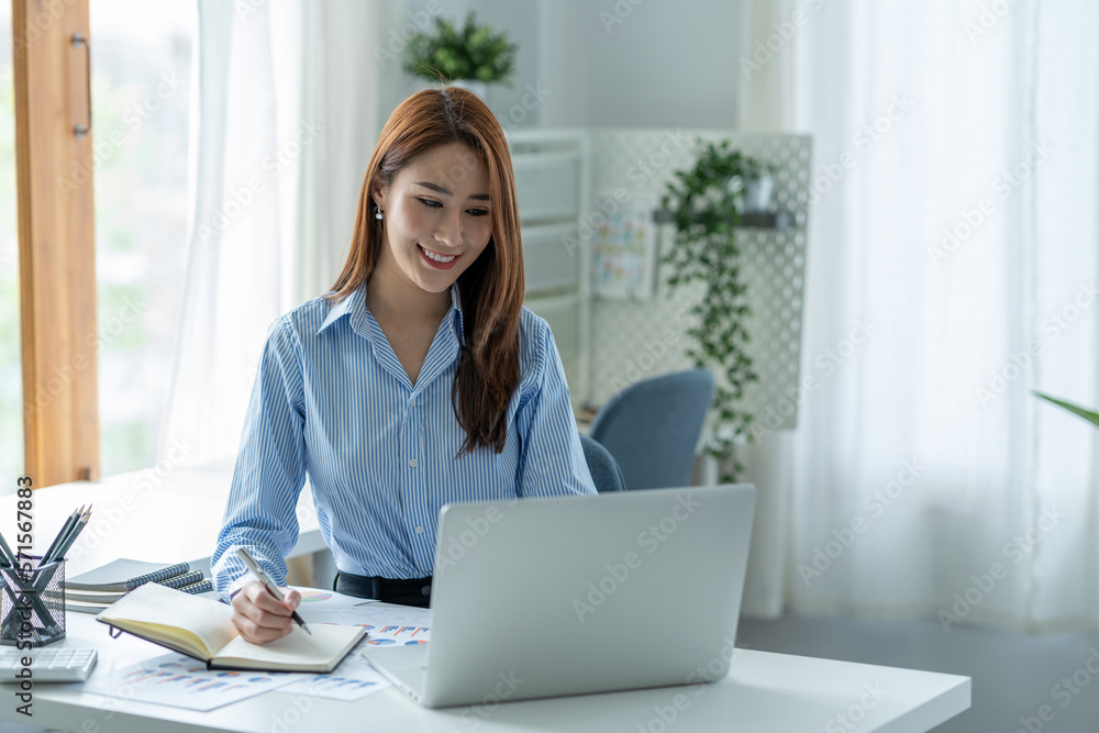 Confident Asian businesswoman sitting and taking notes in financial book Income tax with laptop computer or tablet in a happy office. Confident Asian businesswoman sitting and taking notes in financial book Income tax with laptop computer or tablet in a happy office.