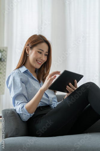 A pretty smiling Asian businesswoman sitting happily holding a tablet on the sofa looking at the desired product details in an online shopping cart in the office.