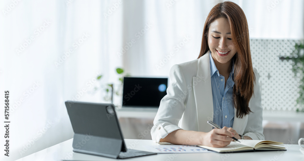 Confident Asian businesswoman sitting and taking notes in financial book Income tax with laptop computer or tablet in a happy office.