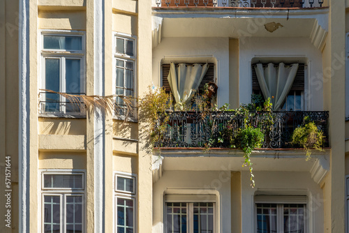 The facade of typical Spanish old house with beautiful balcony with various green plants in sunny winter day. Balcony with curtains and metal railings.