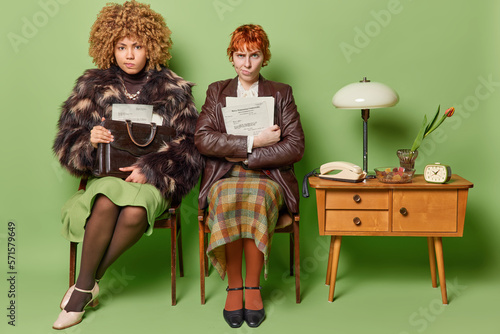 Two women with displeased expression dressed in old fashionable clothes carry papers and antique bag pose on comfortable chairs near table with lamp telephone and clock isolated over green background