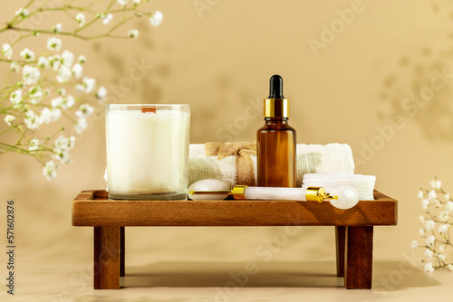 Eco friendly spa relax composition with serum, face roller, towel and candle on wooden podium on beige background with white flowers. Wellness and skin care treatment. Concept of me time and self care