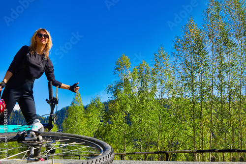 Fototapeta Naklejka Na Ścianę i Meble -  Cute blonde girl in sunglasses stands on road next to lying bicycle, voting with her hand on road. Against background of blue cloudless sky, a young woman athlete stops passers-by with her hand