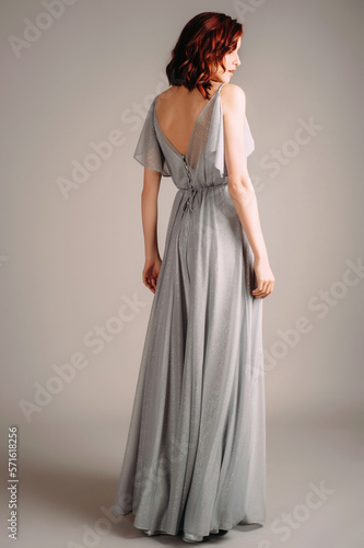 Beautiful young woman in dust blue dress. Studio portrait of ginger lady in evening gown dancing on high heel shoes in studio. Bridesmaid's outfit for hen party, rear view.