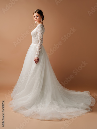 Luxury wedding dress. Fashionable bridal look. Ball gown with tender french lace and beads, long sleeves, lush tulle skirt. Beautiful young blonde bride in studio on brown background. Marriage concept