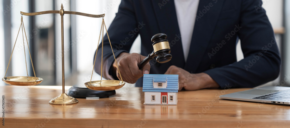 Close-up of a house in front of a lawyer holding a hammer and a laptop silver brass scales on a wooden table in his office, law, legal services, advice, justice and real estate ideas.