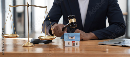 Close-up of a house in front of a lawyer holding a hammer and a laptop silver brass scales on a wooden table in his office, law, legal services, advice, justice and real estate ideas.