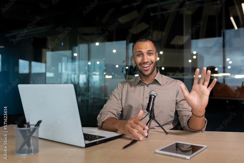 Successful hispanic businessman smiling and looking at camera, portrait of satisfied man in middle of office using microphone and laptop for video call, waving hand in greeting gesture. Successful hispanic businessman smiling and looking at camera, portrait of satisfied man in middle of office using microphone and laptop for video call, waving hand in greeting gesture.