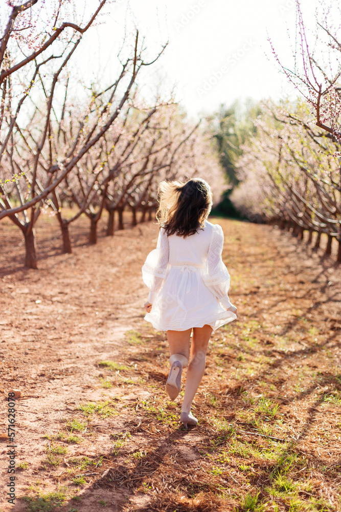 A young beautiful slender girl in a light short dress runs through a blooming peach garden on a sunny spring day. Spring background with a girl in flowering trees A young beautiful slender girl in a light short dress runs through a blooming peach garden on a sunny spring day. Spring background with a girl in flowering trees