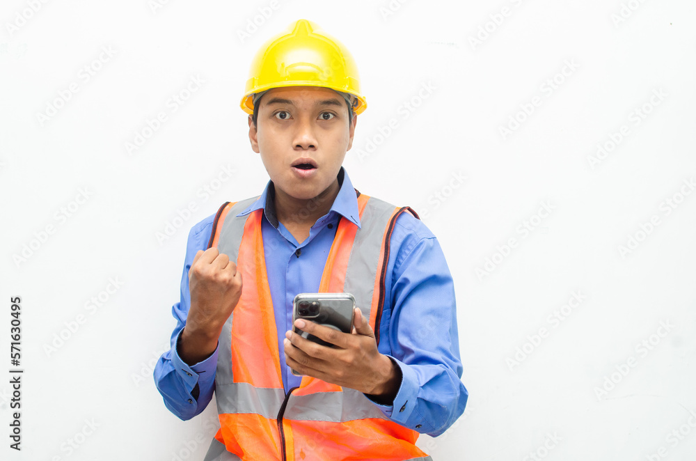 attractive young Asian male construction worker or engineer wearing an orange safety vest and a yellow hardhat holding a phone with shocked, surprised, and amazed expressions. attractive young Asian male construction worker or engineer wearing an orange safety vest and a yellow hardhat holding a phone with shocked, surprised, and amazed expressions.