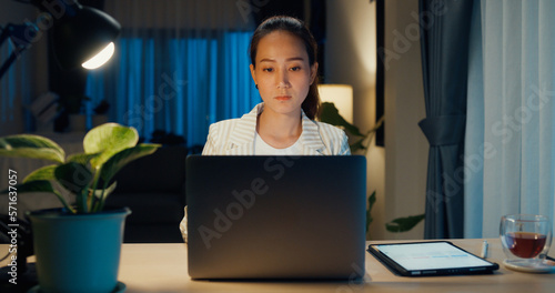 Young Asian businesswoman wear formal sit front of desk with laptop computer and cup of tea focus on screen check report meeting in cozy living room indoor plant at home night. Overtime work concept.