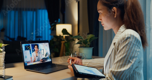 Young Asian businesswoman wear formal sit in front of desk with tablet hold stylus focus on laptop screen video online meeting with colleague in cozy living room at home night. Overtime work concept.