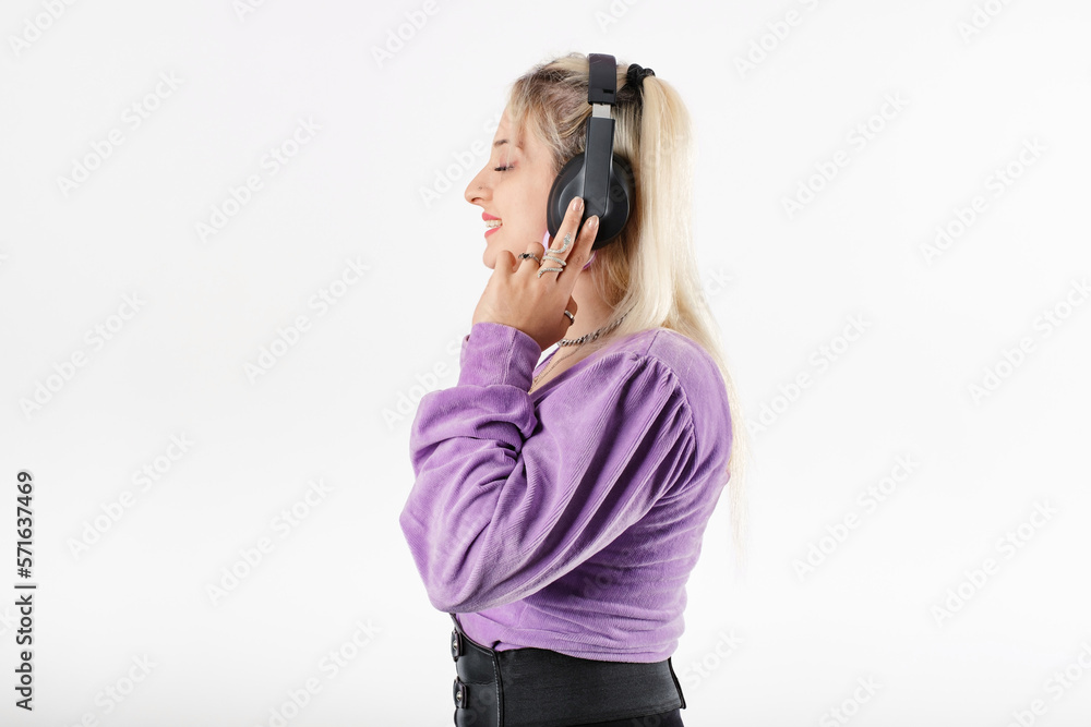 Side view of cute millennial woman wearing blouse standing isolated over white background touching headphones, smiles and looks away and listens to favorite music or podcast. Side view of cute millennial woman wearing blouse standing isolated over white background touching headphones, smiles and looks away and listens to favorite music or podcast.