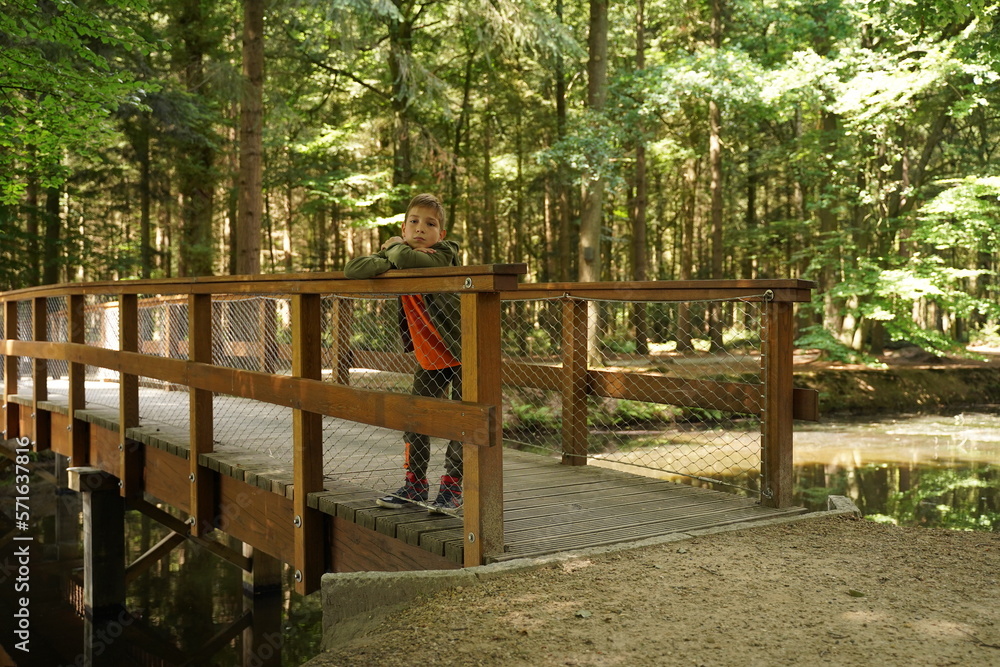 A cute guy of 9-10-11 years old leans on the handrail of a wooden bridge in the forest and looks into the camera. Children's independence, children's loneliness