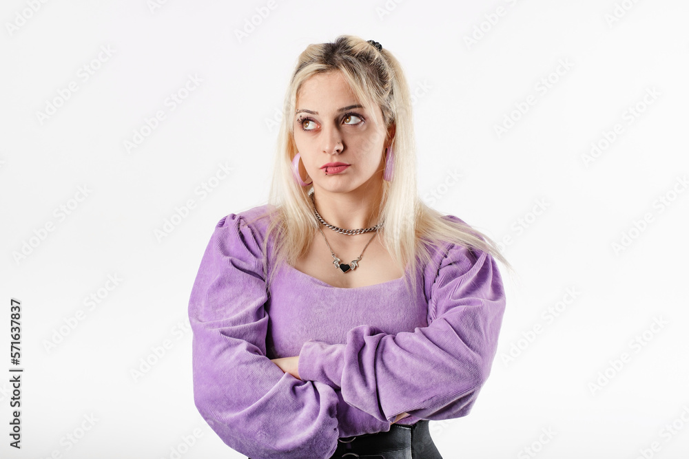 Portrait of young blonde woman wearing ribbed blouse standing isolated over white background with folded arms looks at the empty copy space with an expression of disapproval.