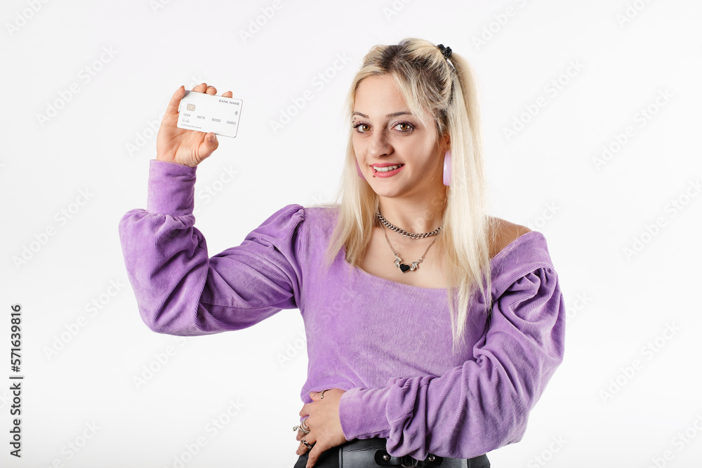 Young blonde woman wearing lilac ribbed blouse isolated over white background holds up credit card and presents it to the camera. Looks at the credit card, the empty copy space, and smiles. Young blonde woman wearing lilac ribbed blouse isolated over white background holds up credit card and presents it to the camera. Looks at the credit card, the empty copy space, and smiles.