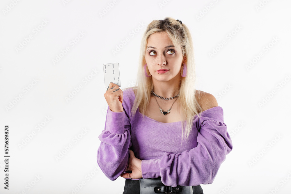 Portrait of cute blonde woman standing isolated over white background holding a credit card looks up with a thoughtful expression. Trying to decide what to buy. Credit card advertising concept. Portrait of cute blonde woman standing isolated over white background holding a credit card looks up with a thoughtful expression. Trying to decide what to buy. Credit card advertising concept.
