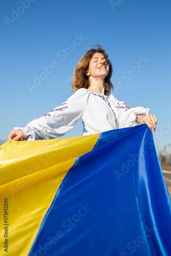 young happy woman in traditional embroidered shirt with yellow and blue Ukrainian flag in foreground. Pride of nation, national identity, faith in victory, symbol of country. independence day, © Anna