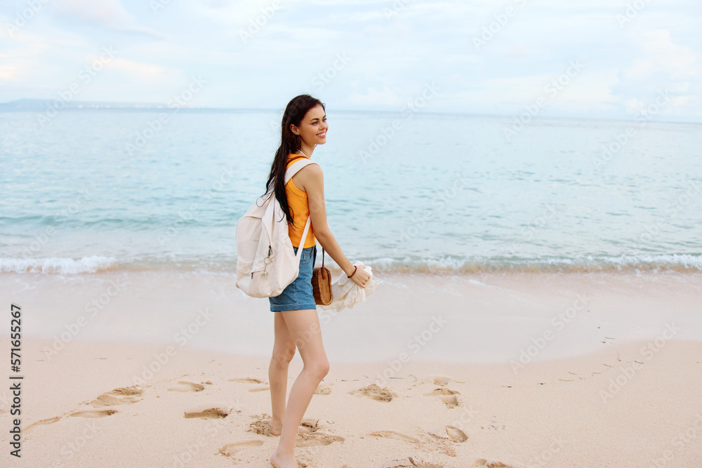 Young woman smile with teeth after swimming in the ocean with a backpack in wet clothes walking along the beach, summer vacation on the island by the ocean in Bali sunset