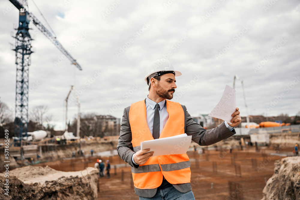 Portrait of a site manager standing on construction site and checking on paperwork and blueprints.