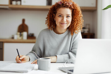 Home portrait of pretty female English tutor working at home sitting at kitchen table, preparing for online lesson with her student, writing in her copybook in front of laptop and cup of coffee