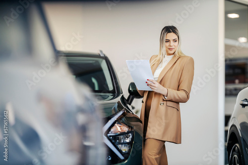 A car dealer is standing at showroom with paperwork in her hands and looking at the cars. © dusanpetkovic1